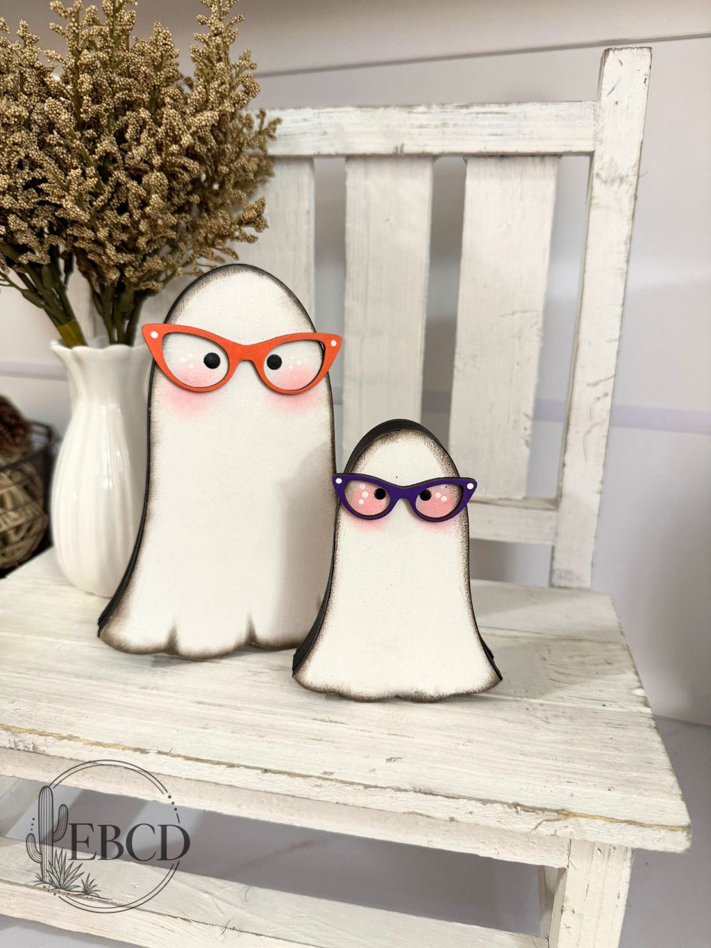 Two ghost-shaped wooden decorations with black edges, one wearing orange glasses and the other purple glasses.