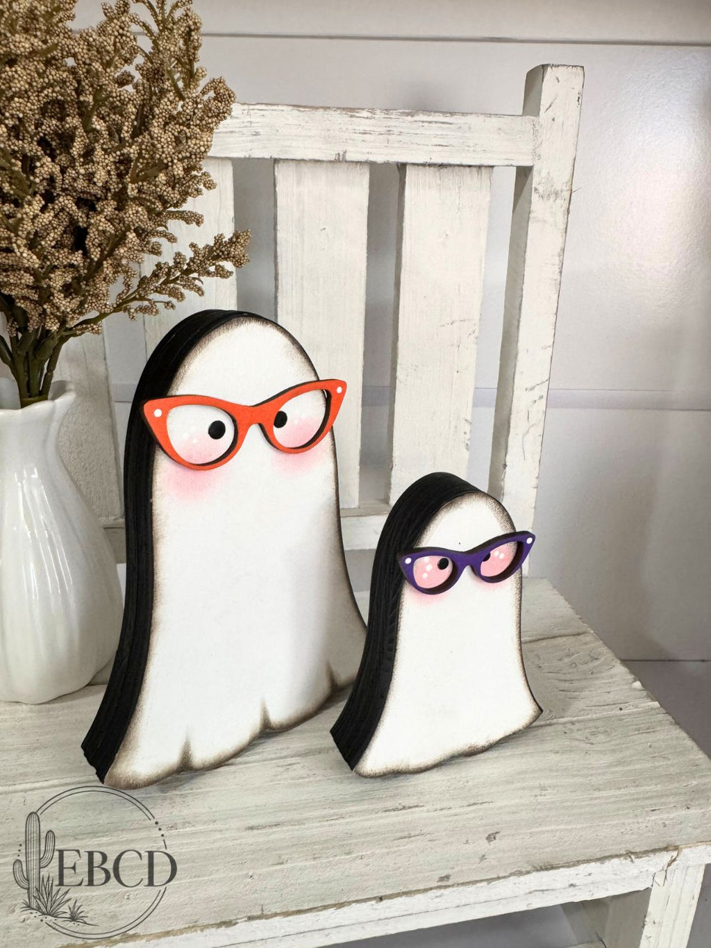 Two ghost-shaped wooden decorations with black edges, one wearing orange glasses and the other purple glasses.