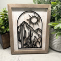 Wooden framed cutout of a desert scene with mountains, cacti, and a sun, placed on a marble surface with plants around.