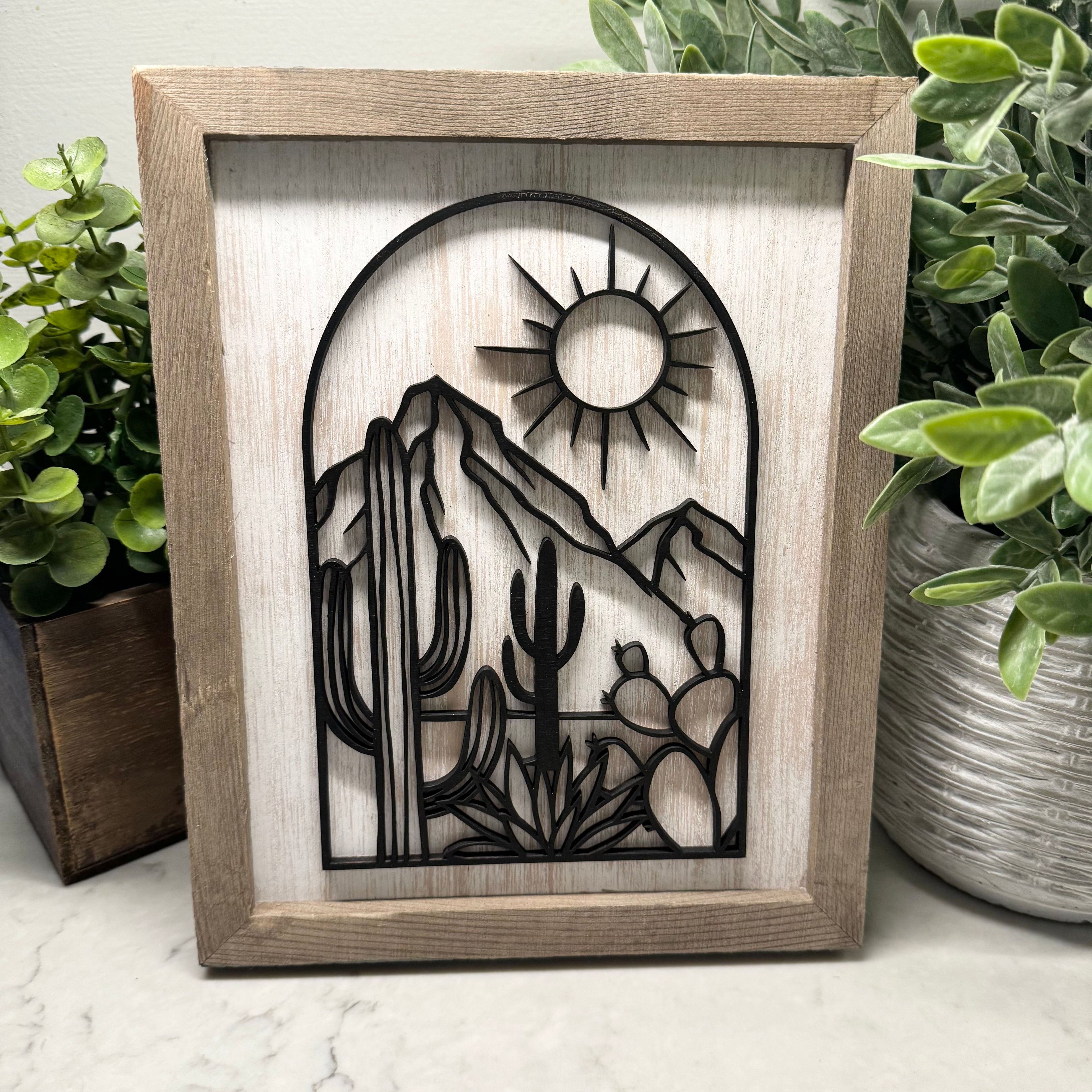 Wooden framed cutout of a desert scene with mountains, cacti, and a sun, placed on a marble surface with plants around.
